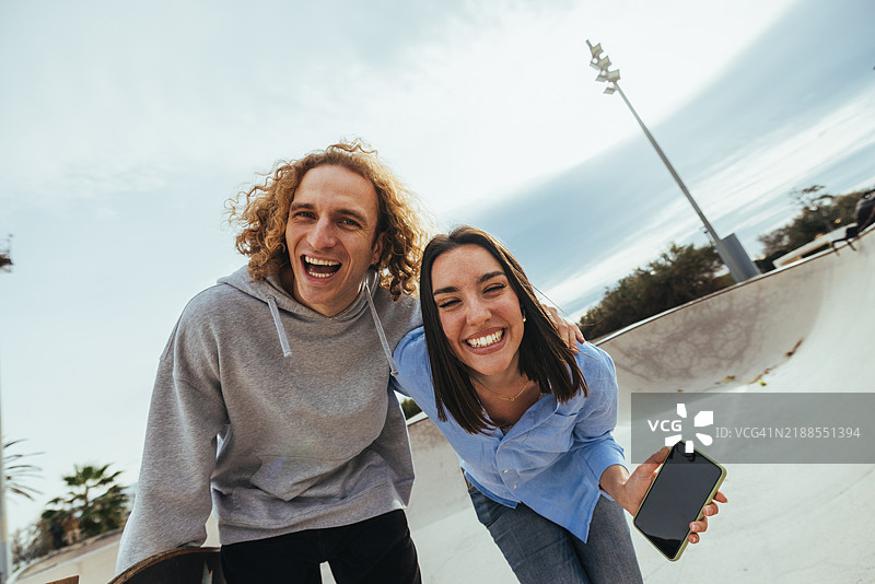 年轻情侣在滑板公园里一起大笑，展示手机。图片素材