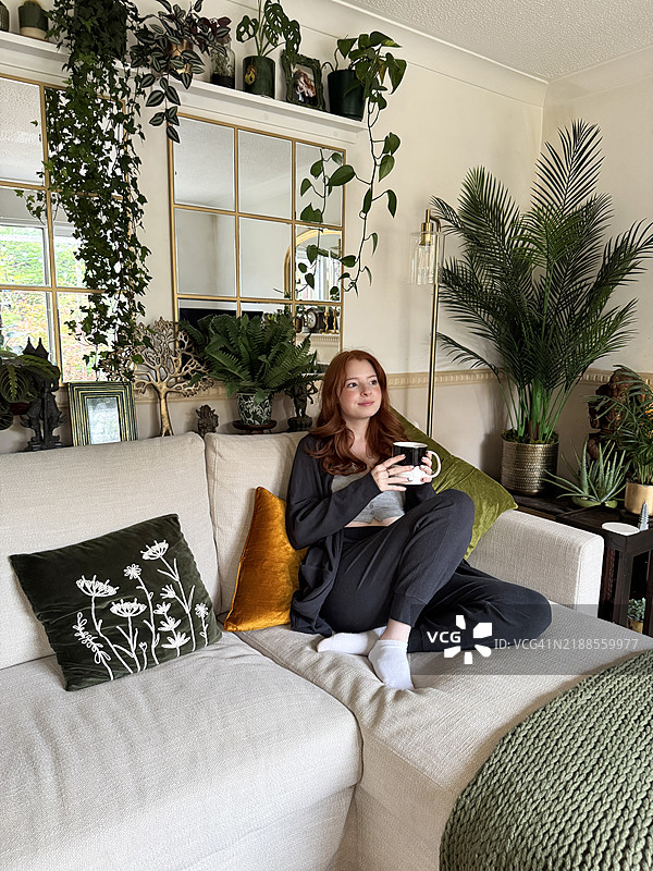 一位迷人的红发年轻女性穿着休闲服，手持黑白咖啡杯，懒散地坐在米色L型沙发上，沙发上有绿色和橙色的天鹅绒靠垫，背景为家庭客厅环境，重点在前景。图片素材