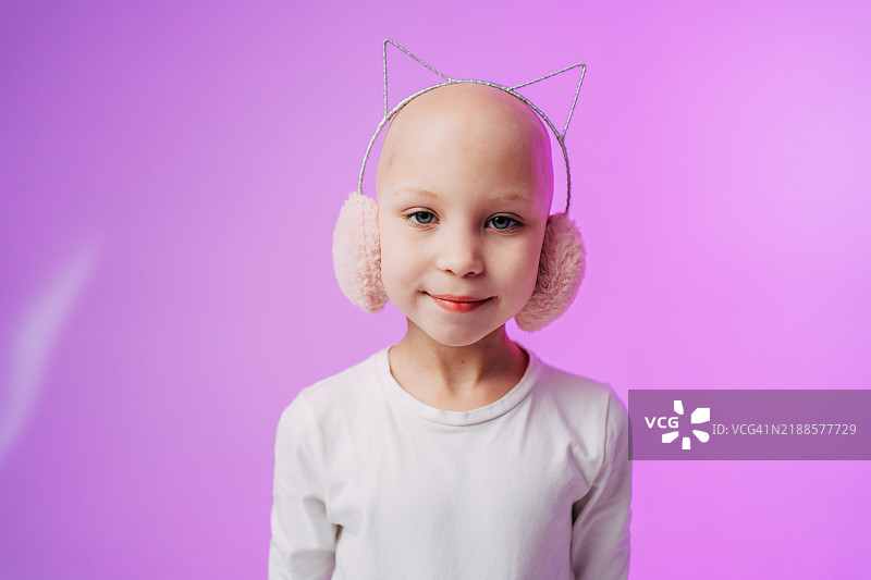 一位微笑的年轻光头女孩，戴着猫耳耳机，背景是鲜艳的紫色。图片素材
