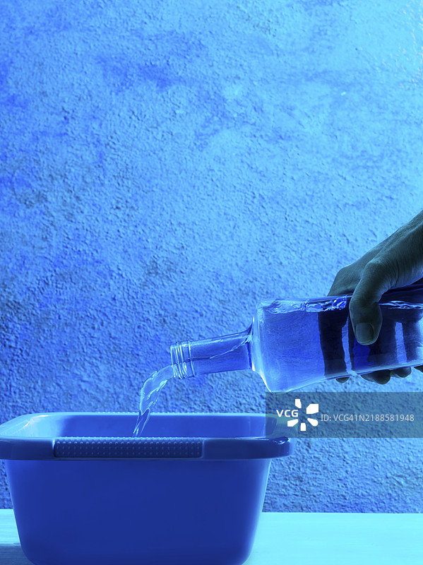 蓝色玻璃瓶倒水，手在全蓝单色的桶和墙上。图片素材