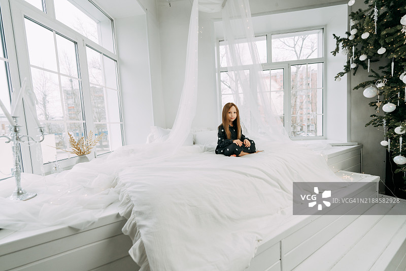 孤独的女孩穿着睡衣坐在圣诞装饰明亮的房间里。图片素材