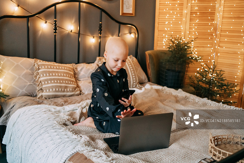 小女孩因脱发而光秃秃的，坐在床上，使用笔记本电脑和智能手机，周围是温馨的圣诞装饰。图片素材