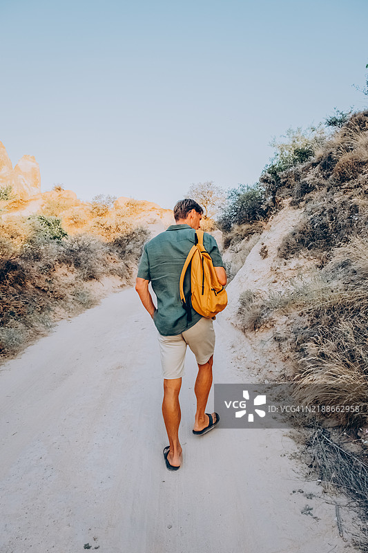 阳光明媚的户外小径上的独自旅行冒险。身穿绿色衬衫、背着背包的帅气男士在卡帕多奇亚山脉徒步旅行。图片素材