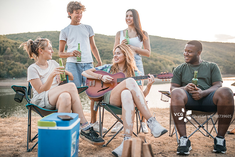 一位快乐的白癜风女性在与朋友露营时弹吉他。他们在湖边享受聚会和喝啤酒的时光。图片素材