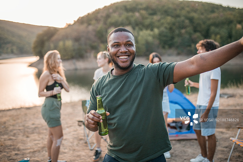 一位英俊的黑人男子在与朋友露营时尽情享乐。他手里拿着一瓶啤酒，正在跳舞。图片素材