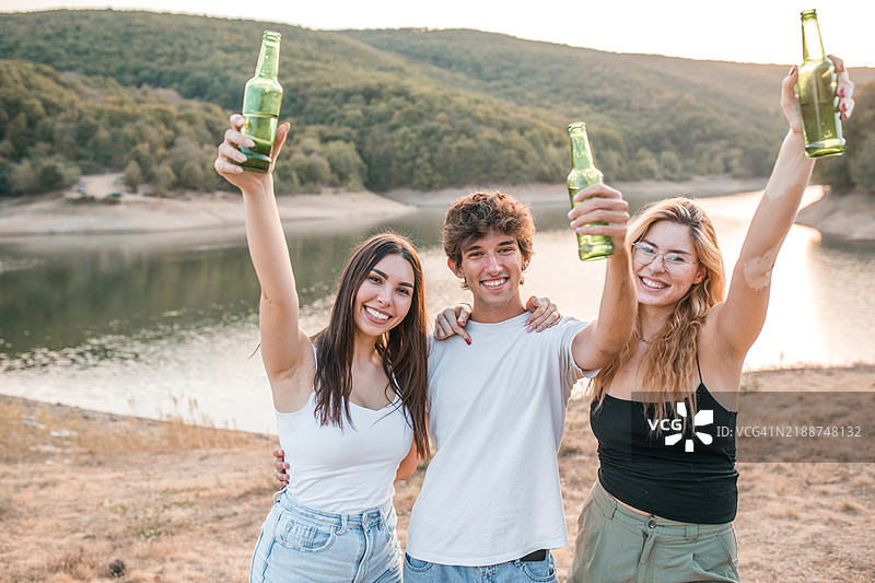 三位朋友站在湖边，举起啤酒瓶，直视镜头。图片素材