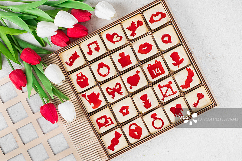 可爱的方形饼干，里面夹有红色果酱，造型为情人节主题的图案。放在一个木制礼盒中，装饰有郁金香。白色背景。俯视图。图片素材