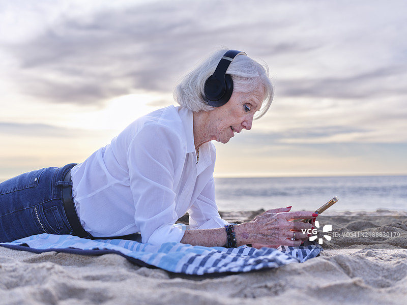 一位白发女士在海滩上听着应用程序里的音乐，同时发送WhatsApp消息。图片素材