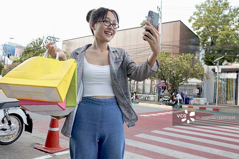 亚洲年轻女商人手持购物袋，边走过城市街道的斑马线边用手机自拍。女性工作人员在户外办公室使用智能手机。快乐的女性在现代城市户外使用电子设备。图片素材