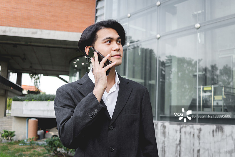 身穿黑色西装的亚洲年轻商人在现代建筑前打电话。男性工作人员在户外办公室使用智能手机。快乐的聪明商人在城市现代环境中使用电子设备。图片素材