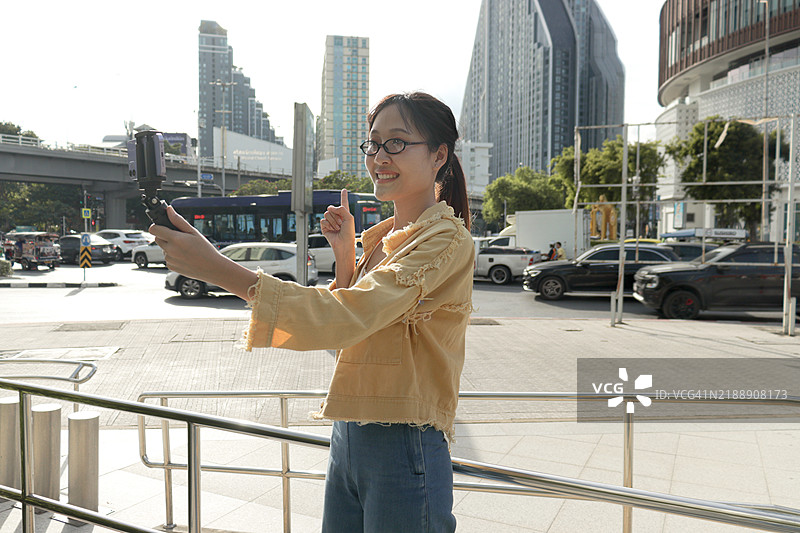 亚洲年轻女性在城市人行道上看着手机并自拍，背景是现代建筑。女性在城市中使用智能手机。快乐的女性在现代城市环境中户外使用电子设备。图片素材