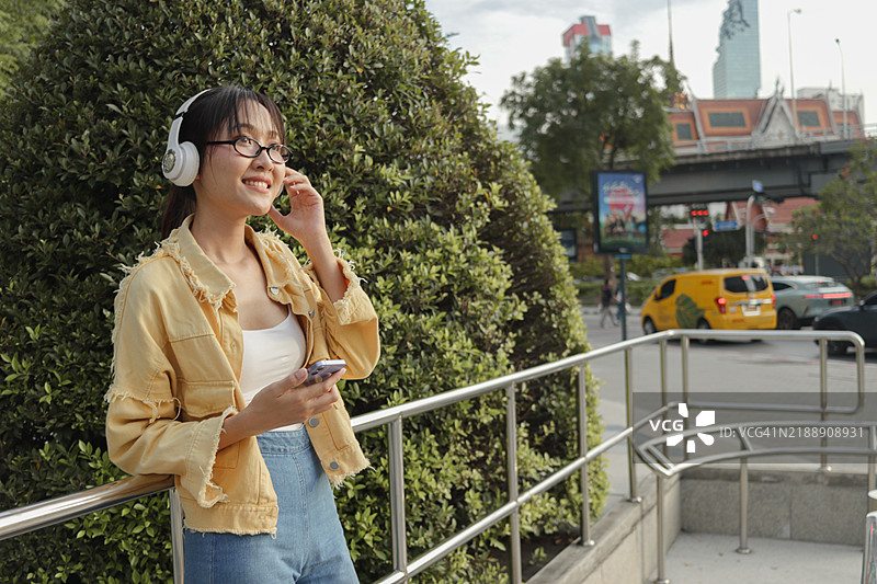 亚洲年轻女性在城市人行道上站着，手持手机，戴着耳机听音乐，背景是现代建筑。女性在城市中使用智能手机。快乐的女性在现代城市环境中户外使用电子设备。图片素材