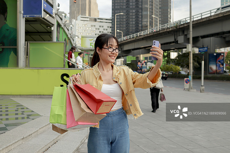 年轻的亚洲女性手持购物袋，站在人行道上，望着手机自拍，背景是现代建筑。女性在城市中使用智能手机。快乐的女性在现代城市环境中户外使用电子设备。图片素材