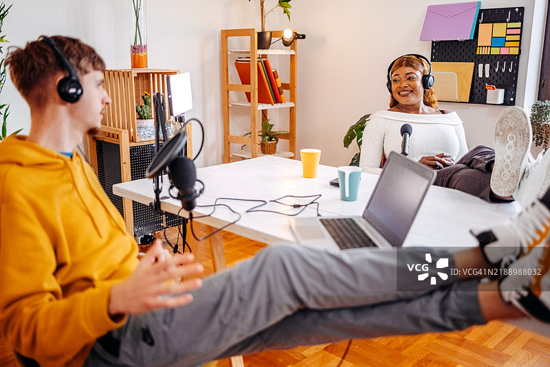 年轻人在家中轻松地坐着，脚抬起，愉快地拍摄播客，面对着摄像机交谈。图片素材