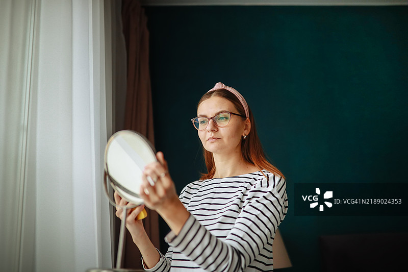 舒适的造型时刻：一位女性在绿色墙壁的卧室里拿着镜子，试戴眼镜和头带，将在线购物与居家便利相结合。图片素材