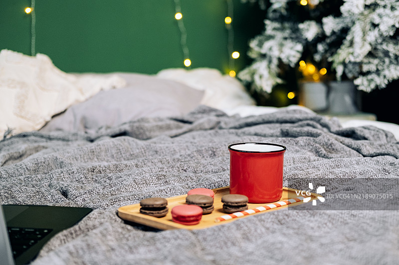 红色咖啡杯和马卡龙放在木托盘上，背景是绿色墙壁和圣诞花环，摆放在床上。图片素材