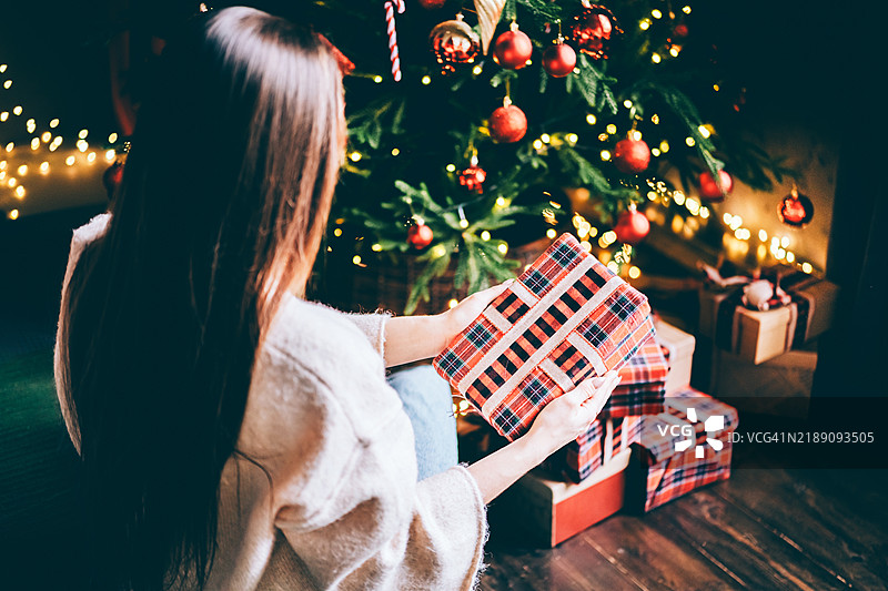 一位面容模糊的女性穿着柔软的羊毛针织毛衣，将包裹好的礼物盒放在明亮的圣诞树下。图片素材
