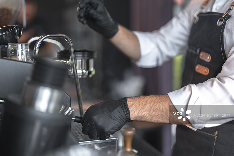 一位穿着围裙、戴着手套的咖啡师在浓缩咖啡机前准备咖啡，重点展示手部和设备。图片素材