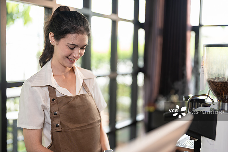 一位微笑的咖啡师穿着白衬衫和棕色围裙，在咖啡馆中操作咖啡研磨机，阳光透过大窗户洒入室内。图片素材