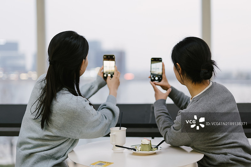两个女孩在咖啡馆享受休息，使用手机拍摄外面的风景。图片素材