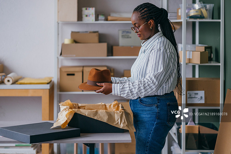 女性在小型商业工作空间中包装帽子，周围有货架。图片素材