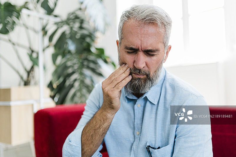 老年男性正感到不适，背部疼痛，坐在家中客厅的沙发上按摩酸痛的肌肉。老年人和健康问题的概念图片素材