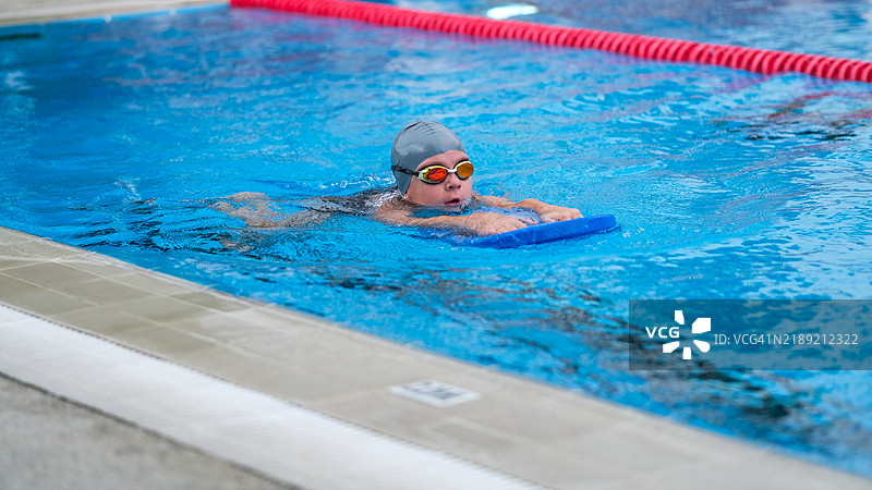 一个年轻女孩在明亮的蓝色泳池中用浮板练习游泳。图片素材