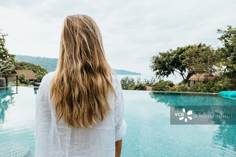 长发女人站在无边泳池旁，面朝大自然。图片素材