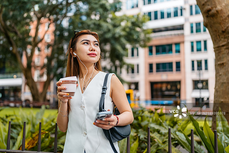 年轻亚洲女性在澳大利亚悉尼的街道上喝咖啡、听音乐，同时使用手机。图片素材