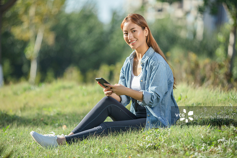 一位年轻的亚洲女性坐在草地上使用手机图片素材