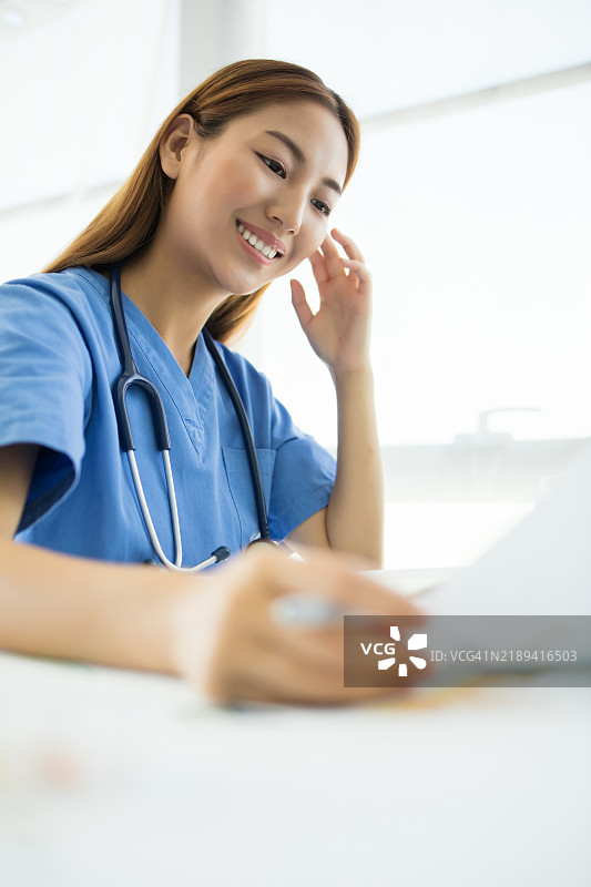 穿着蓝色制服、脖子上挂着听诊器的亚洲女性医生或护士，面带微笑，手持夹板正在写字或阅读。图片素材