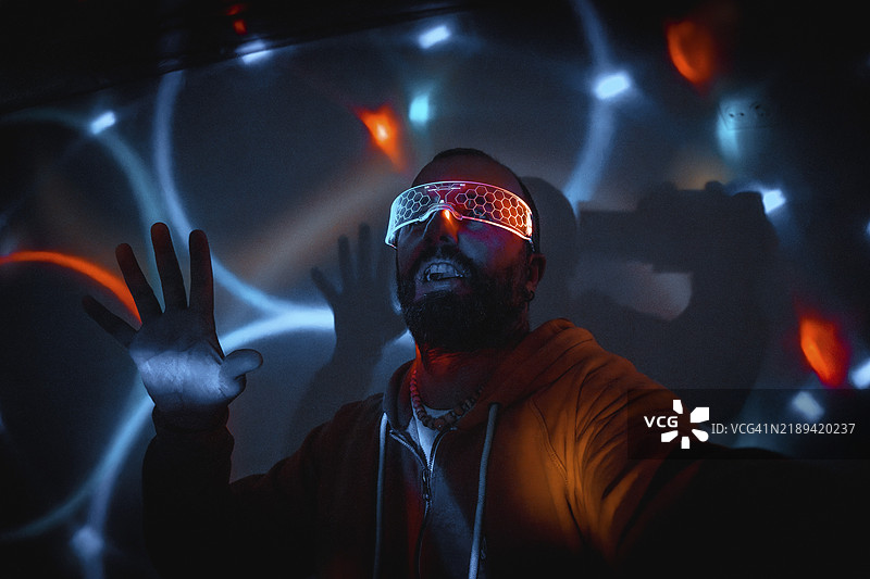 戴着未来派眼镜的胡子男人在黑暗的房间里与虚拟现实互动，手部动作和兴奋的表情伴随着五彩斑斓的霓虹灯。图片素材