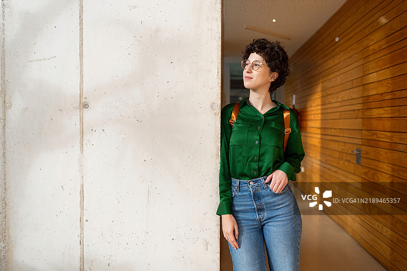 戴眼镜和背包的女商人在可持续现代办公室的混凝土墙上倚靠，沉思中。图片素材