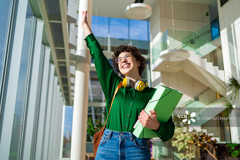 快乐的大学生手持文件夹，在现代环保的大学校园中挥手，阳光充足。图片素材