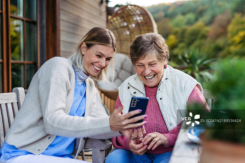 护士和女性老年患者在家中查看手机里的照片。老年护理和家庭护理概念。图片素材