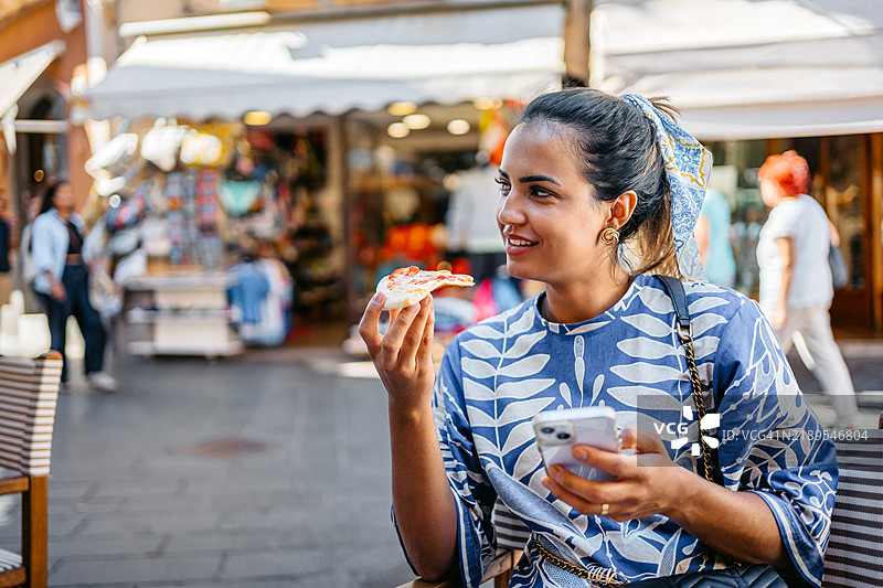 年轻的巴西女性在西西里岛陶尔米纳的一家户外餐厅里吃着一片披萨，同时使用手机。图片素材