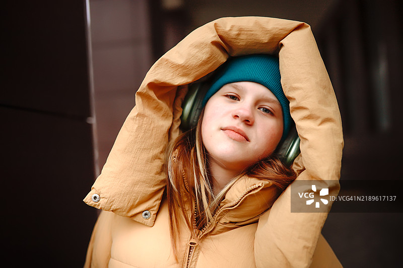 温暖的冬季造型：戴着耳机和连帽外套的少女，拥抱寒冷的城市街道。正看向镜头。图片素材