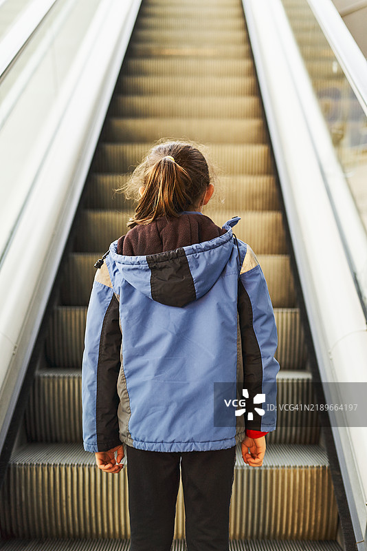 年轻女孩穿着夹克站在购物中心的扶梯上，背影。图片素材