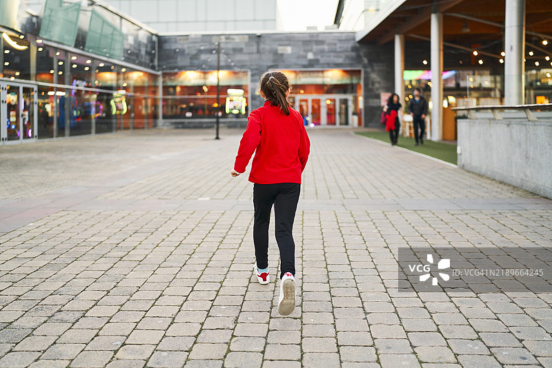 一个穿着红色毛衣的女孩在购物中心的走廊里奔跑，背景中有其他人。图片素材