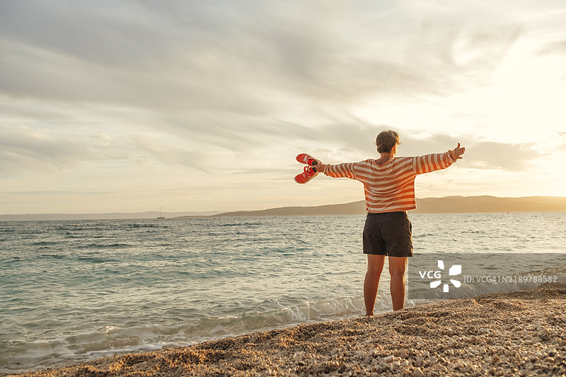 自由快乐的女人赤脚站在海中，手里拿着运动鞋，双臂向两侧伸展，放松并享受这一刻。图片素材