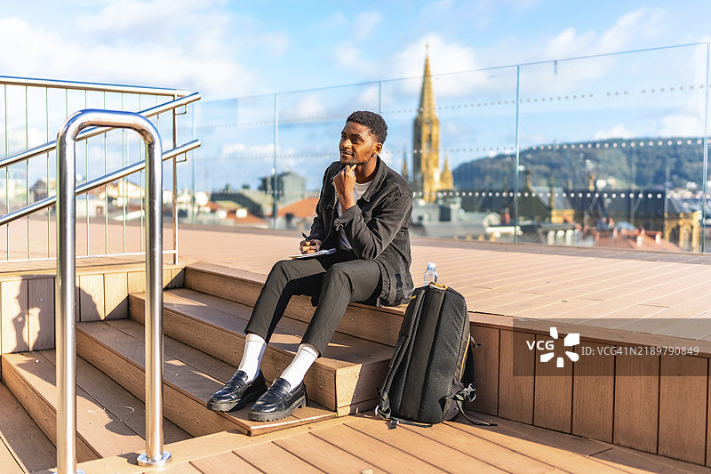 沉思的非洲年轻男性旅行者在阳光明媚的日子里坐在城市观景点写笔记图片素材