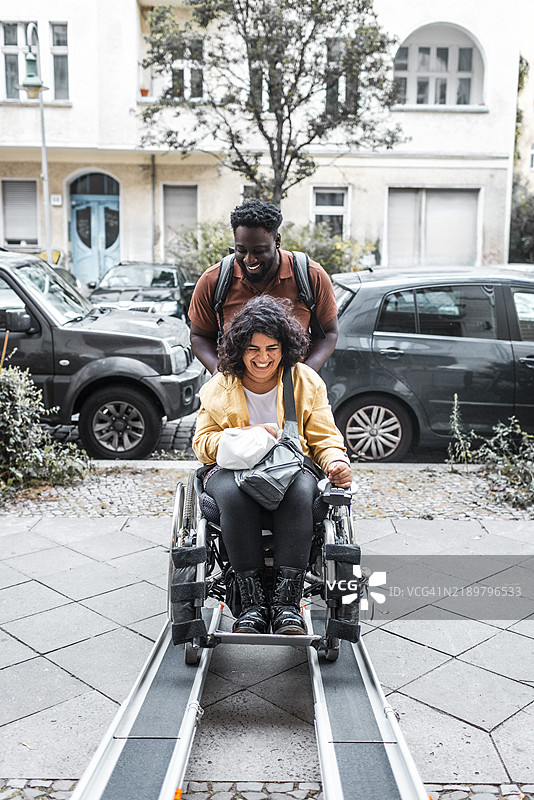 快乐的年轻男子在人行道的坡道上推着坐在轮椅上的女子图片素材