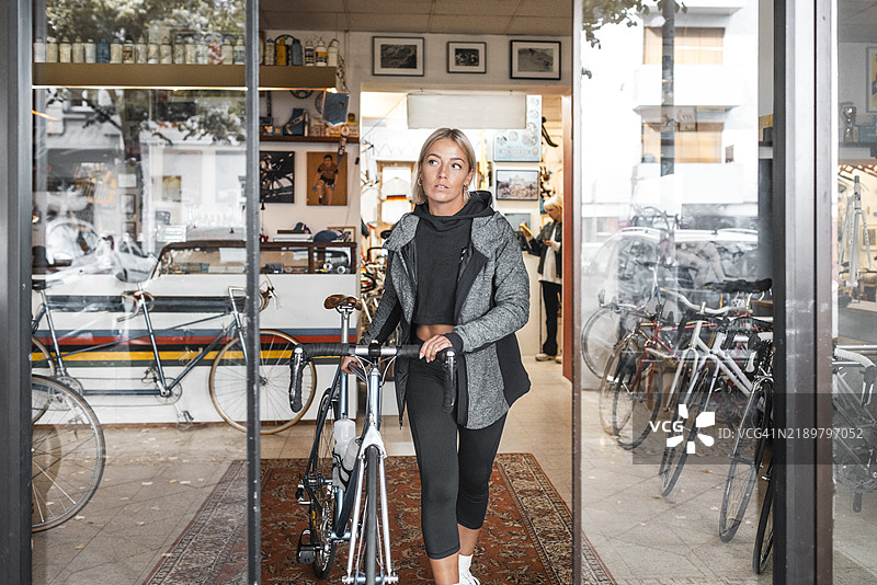 一位深思的女性穿着运动服，骑着自行车从自行车店的门口走出。图片素材