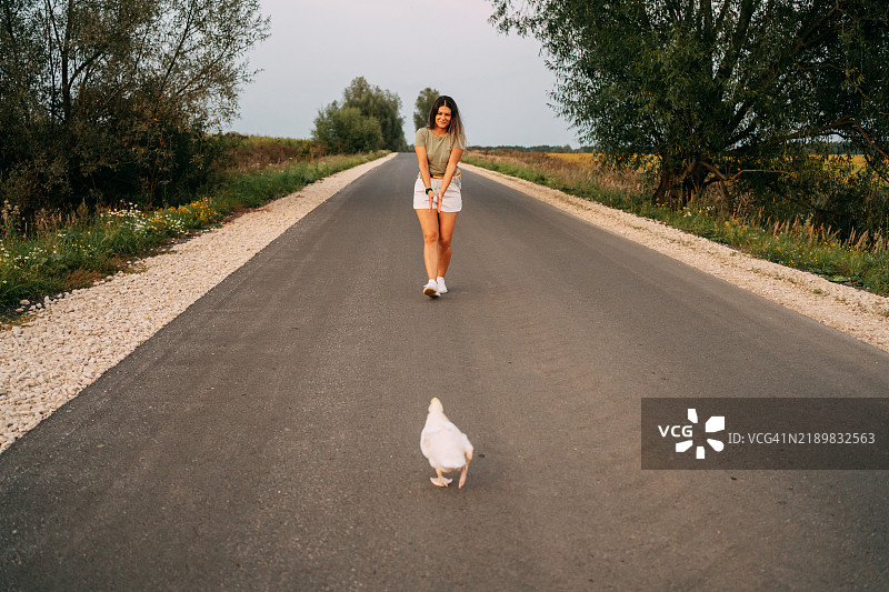 年轻女子沿着铺好的乡村道路走，身后跟着一只白色的鸡。图片素材