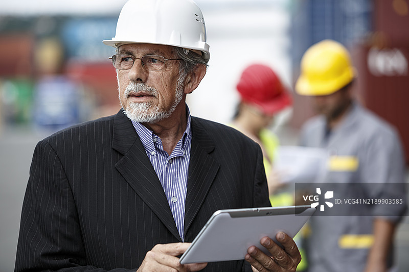 一位自信的工头站在码头上，戴着白色安全帽，使用数字平板电脑监督仓库的运输操作，检查集装箱，管理物流和库存控制。图片素材