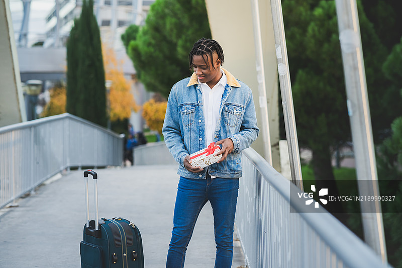 年轻商人拖着行李箱，手里拿着礼物，正看着它。图片素材