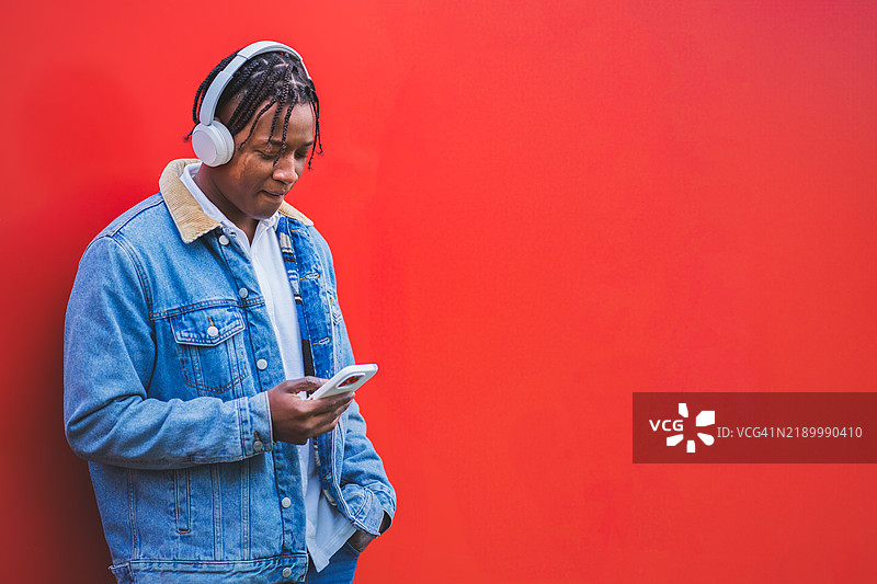 年轻的拉丁美洲男子在红色背景下使用智能手机，戴着耳机。图片素材