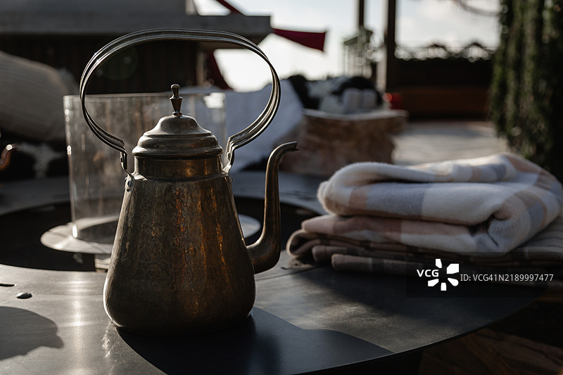 复古茶壶放在深色表面上，背景是模糊的露台和叠好的毛毯。图片素材