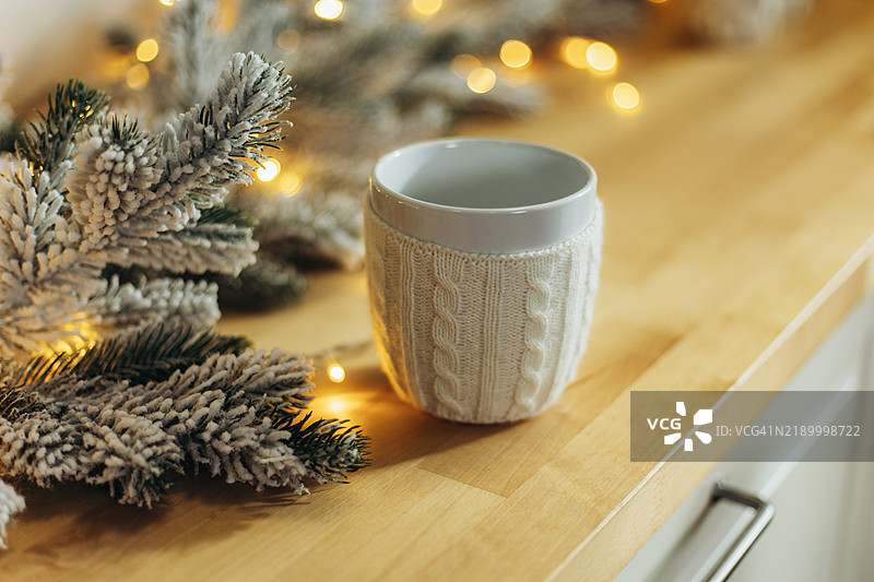 白色马克杯上有针织套，放在木质台面上，旁边是雪白的松枝和串灯。图片素材