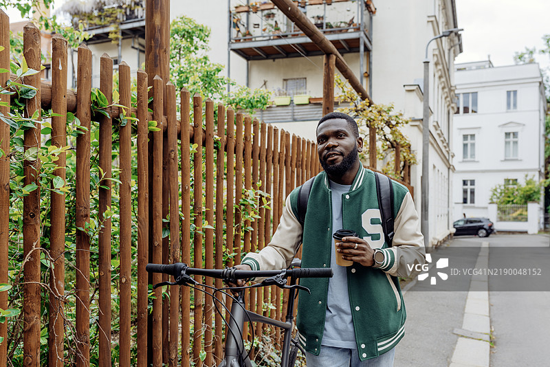 一位穿着绿色夹克和背包的年轻有色人种男子在莱比锡的一条温馨街道上推着他的自行车，手里拿着外带咖啡。图片素材
